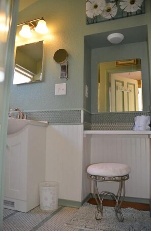 Bathroom features a sink, vanity, and stool with cushioned seat, along with a mirror and decorative flower arrangement.