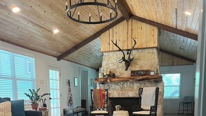 A spacious living room with a stone fireplace, wooden floors, and a high ceiling with exposed beams. - Completely renovated 1930s Cottage on the Guadalupe River. Ideal location! (New Braunfels)