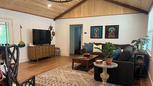 A living room with wooden floors, a TV, a sofa, a coffee table, and a large window. - Completely renovated 1930s Cottage on the Guadalupe River. Ideal location! (New Braunfels)