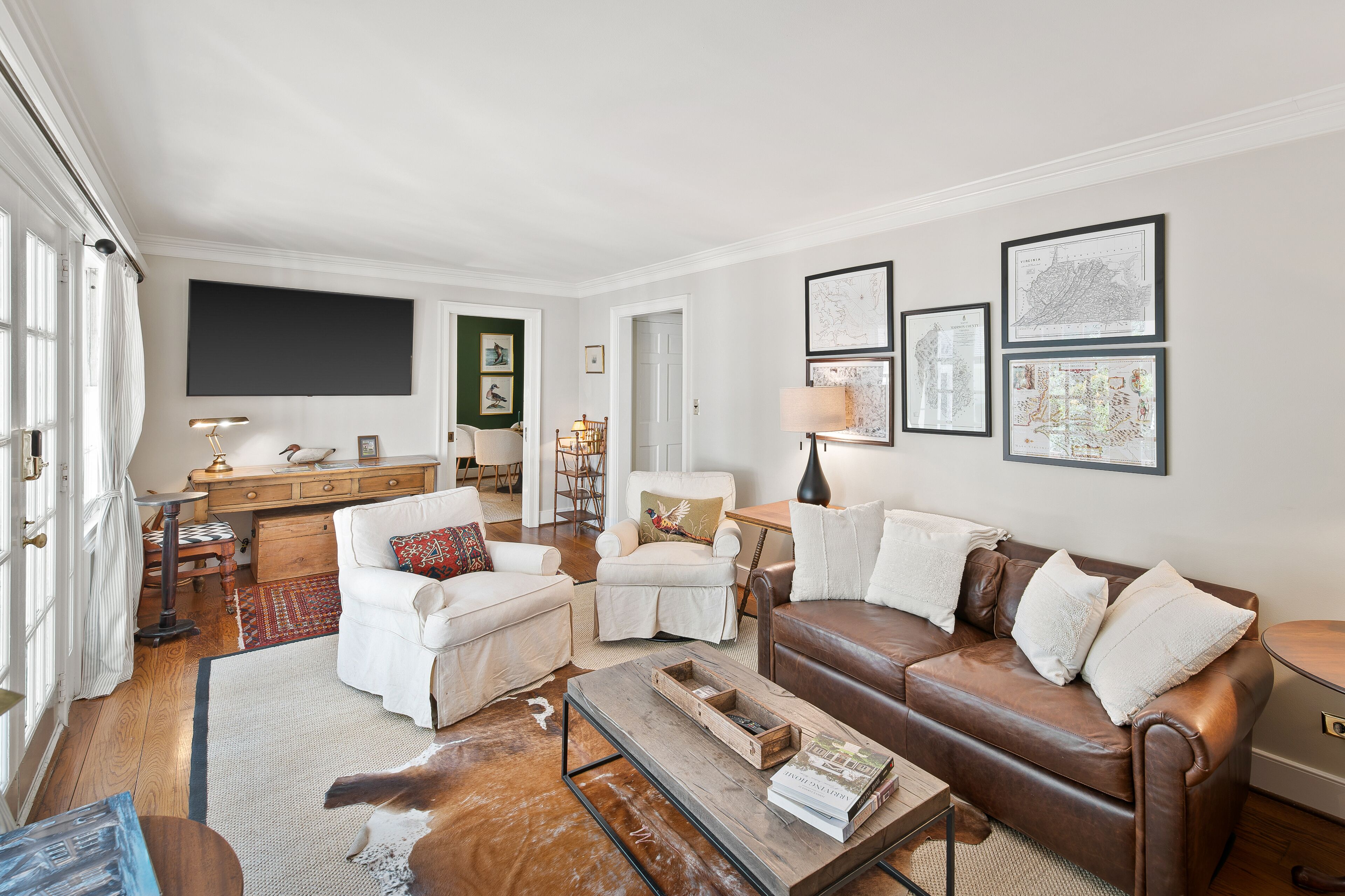 A living room with a brown leather sofa, white armchairs, a coffee table, and a flat-screen TV mounted on the wall.