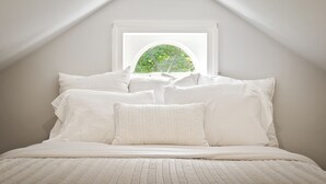 A bedroom with a sloped ceiling, a large window with a view of greenery, and a neatly made bed with white bedding. - GreenLeaf | Close to UVA and Downtown, Sleeps 7 with Yard! (Charlottesville)