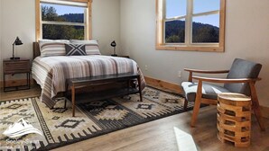 Un dormitorio con cama, dos mesitas de noche, una silla, alfombra y dos ventanas con vistas a árboles y colinas.