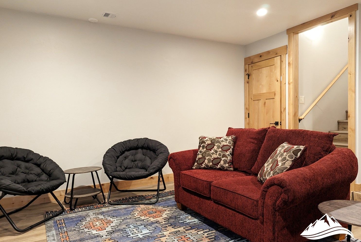 A cozy living room with a red sofa, two black chairs, a patterned rug, and a wooden door.