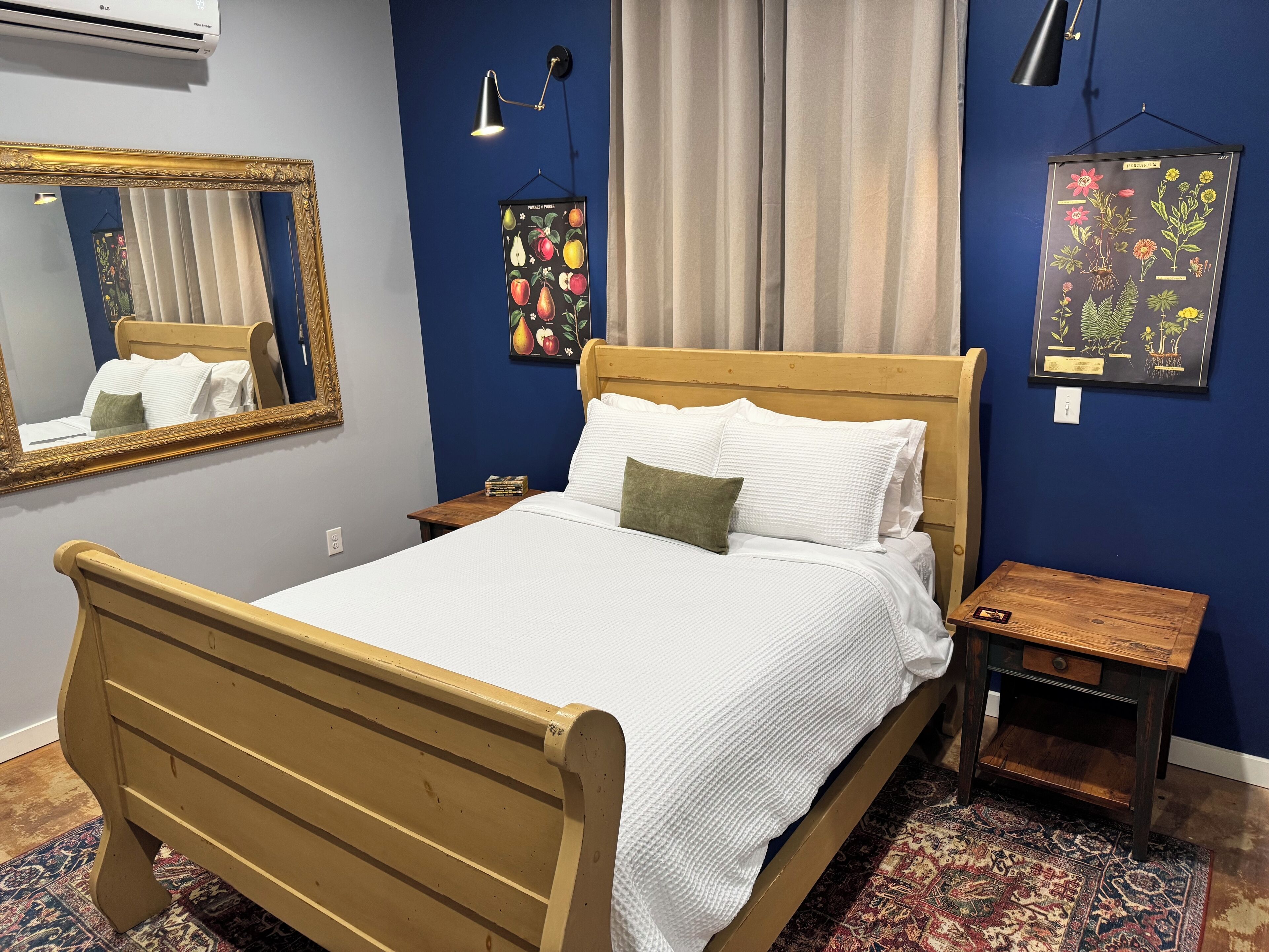 A bedroom with a wooden bed, a nightstand, a mirror, a wall-mounted lamp, and a botanical print.