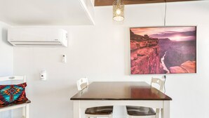 A dining area with a dark wooden table, white chairs, and a colorful cushion. An air conditioner is mounted on the wall, and a framed picture of a canyon is displayed above the table. - The Teal Door | UofA Central Newly Renovated 1BR (Tucson)
