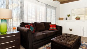 A living room with a brown leather sofa, a matching ottoman, a wooden side table with a lamp, and a patterned rug. - The Teal Door | UofA Central Newly Renovated 1BR (Tucson)