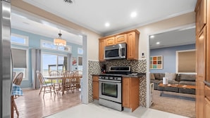 A modern kitchen with wooden cabinets, a microwave, and an oven. There is a dining area with a table and chairs, and a living room with a sofa and a coffee table.