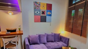 Una sala moderna con un sofá seccional morado, una pequeña mesa de madera con plantas y un cuadro abstracto colorido en la pared.