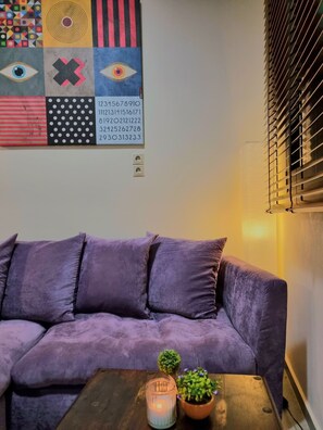 Ein violettes Sofa mit einem kleinen Tisch davor, eine Wand mit einem farbenfrohen, abstrakten Kunstwerk und ein Fenster mit Jalousien.