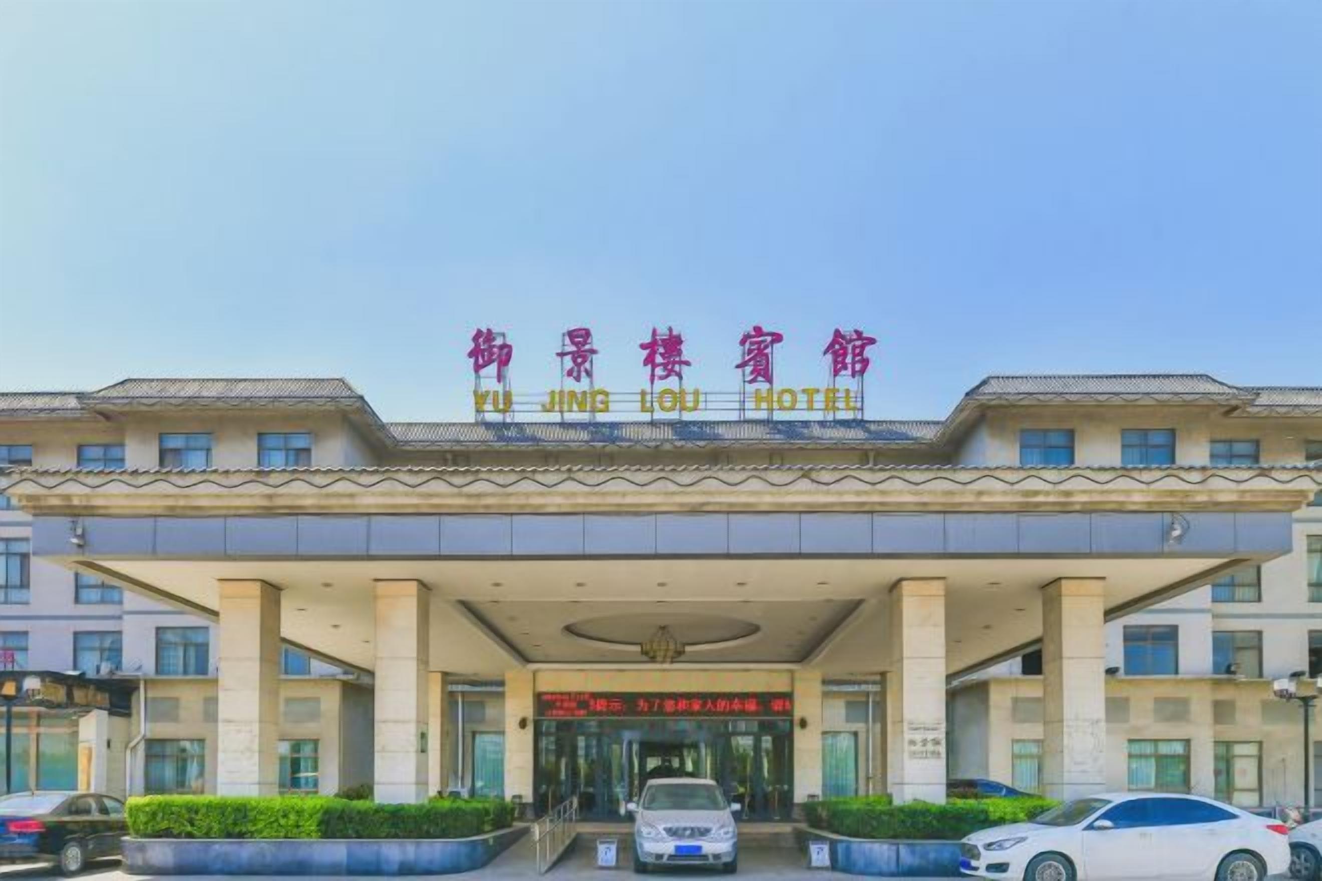 Entrada de un hotel con un letrero en idioma extranjero, una galería techada y varios coches estacionados frente a él.