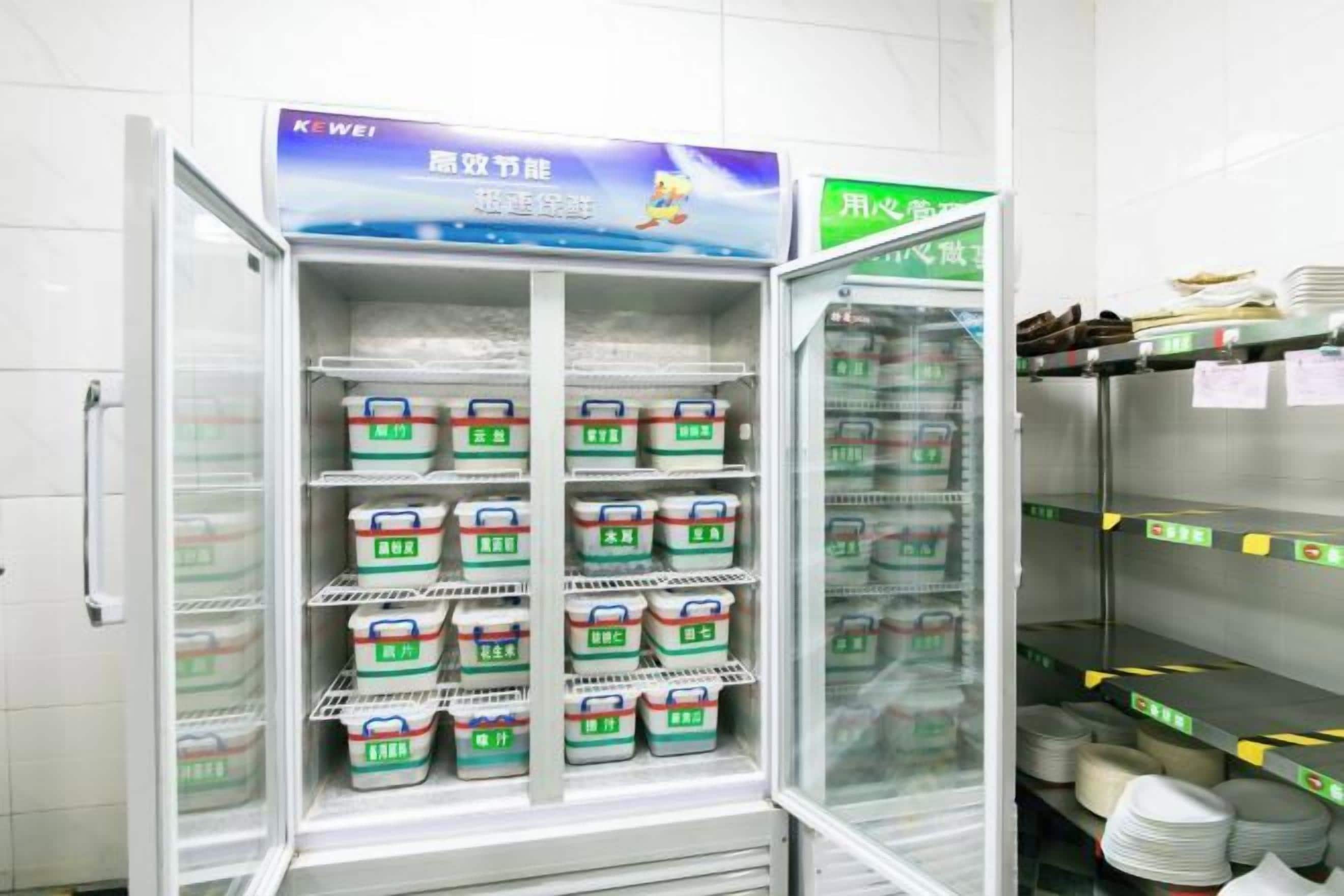 Un refrigerador comercial lleno de alimentos, un compartimento congelador y una estantería con platos y cubiertos.