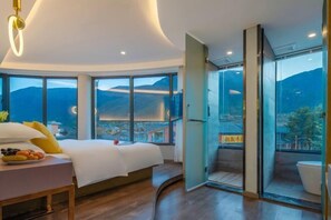 Una habitación de hotel moderna con una cama grande, una mesita de noche con fruta y un vidrio corredizo que da a un balcón con vistas a la montaña.