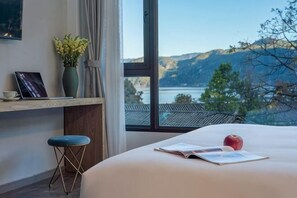 Un dormitorio con un ventanal grande, una cama, un escritorio con silla y vistas a un lago y montañas.