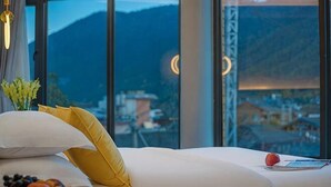 A hotel room with a bed, pillows, a fruit plate, and a view of a mountainous landscape through large windows. - Lugu Lake Yueji Hotel (Ninglang)