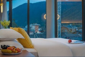 Habitación de hotel con cama, almohadas, una bandeja de frutas y vistas a un paisaje montañoso a través de amplias ventanas.