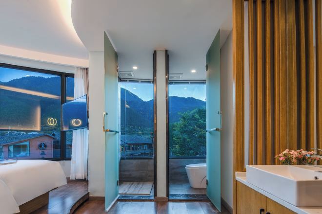 Una habitación de hotel moderna con una cama grande, un baño con lavamanos y vistas a montañas a través de un ventanal.