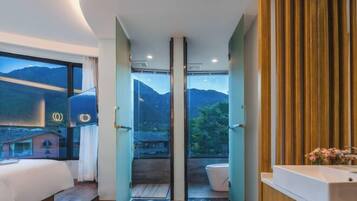 Una habitación de hotel moderna con una cama grande, un baño con lavamanos y vistas a montañas a través de un ventanal.