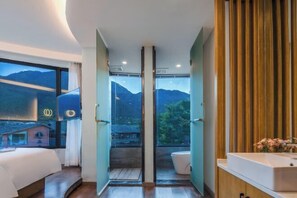 Una habitación de hotel moderna con una cama grande, un baño con lavamanos y vistas a montañas a través de un ventanal.
