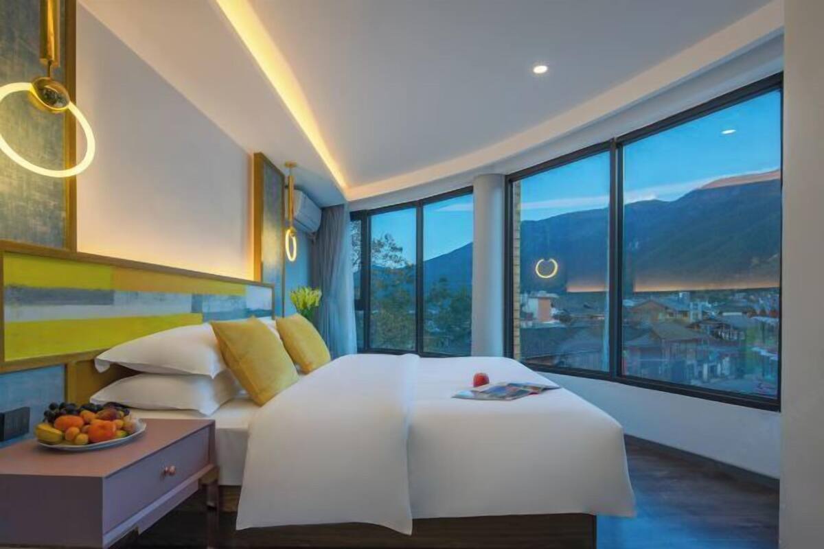 Una habitación de hotel moderna con una cama grande, una mesita de noche con fruta y vistas a la ciudad a través de amplios ventanales.