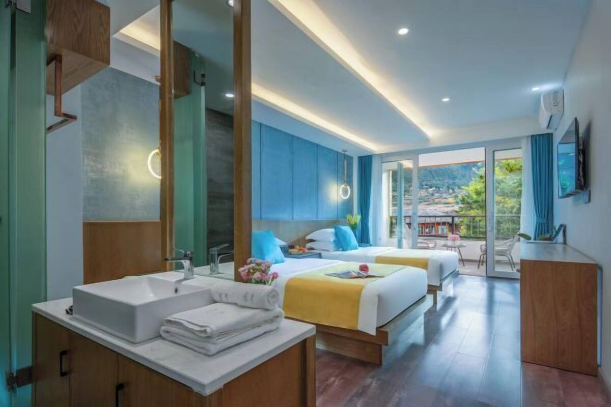 Habitación de hotel moderna con una cama grande, un sofá independiente, un baño con lavamanos y espejo, y un balcón con vistas.