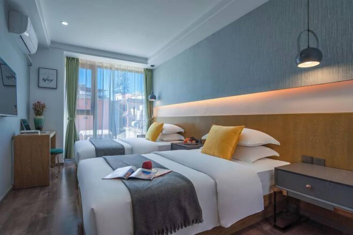 Habitación de hotel con dos camas, un escritorio, una silla, una ventana con cortinas y una lámpara fijada a la pared.