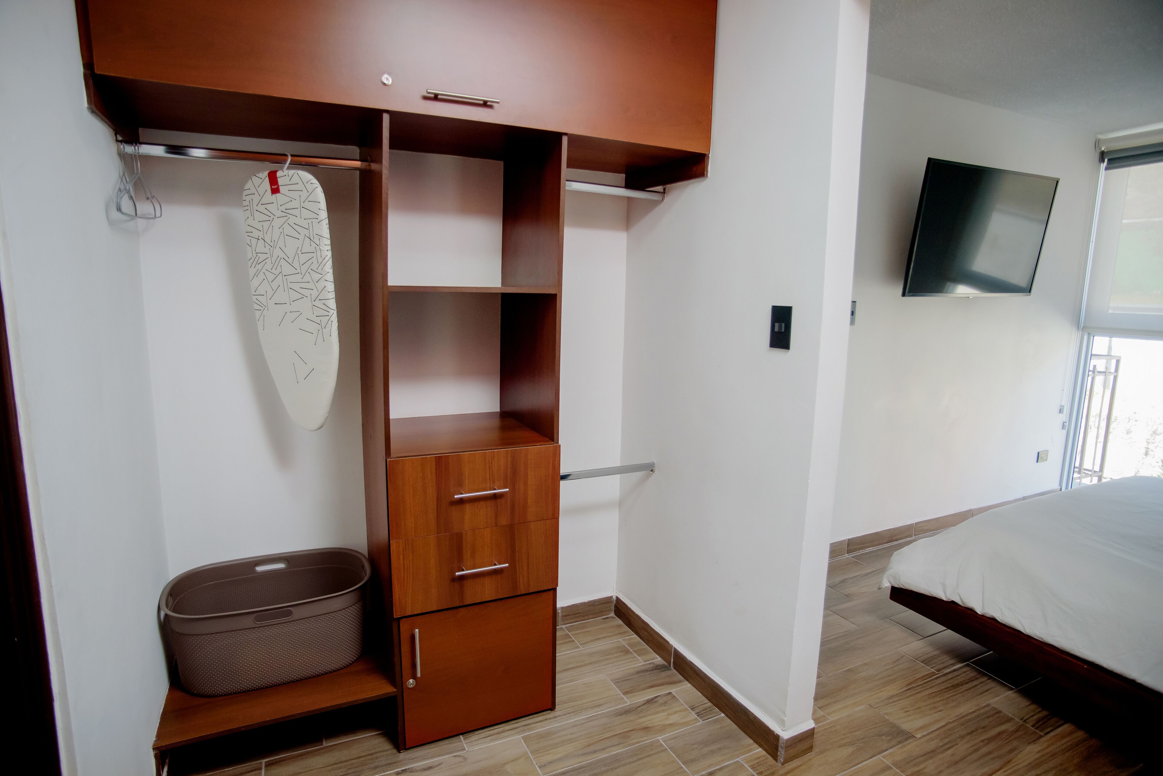 Un dormitorio con cama, armario con varios compartimentos, televisor montado en la pared y ventana que proporciona luz natural.