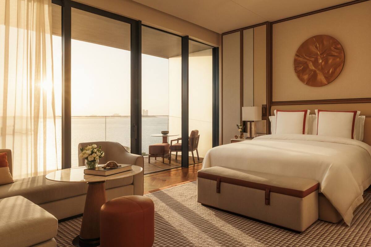 A hotel room with a large bed, a sitting area with a sofa and chairs, a small table with a vase of flowers, and a view of the sea through large windows.