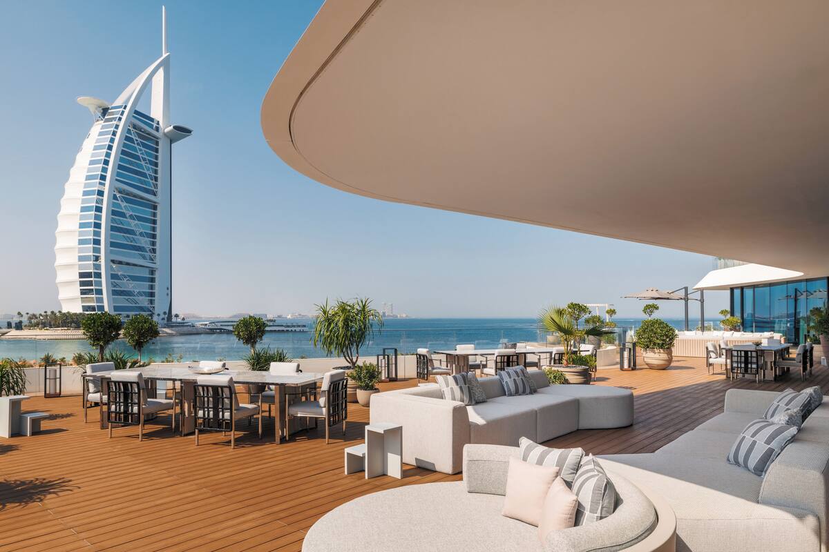 A modern outdoor lounge area with a curved ceiling, furnished with sofas, tables, and chairs, overlooking a body of water and a distinctive sail-shaped building.