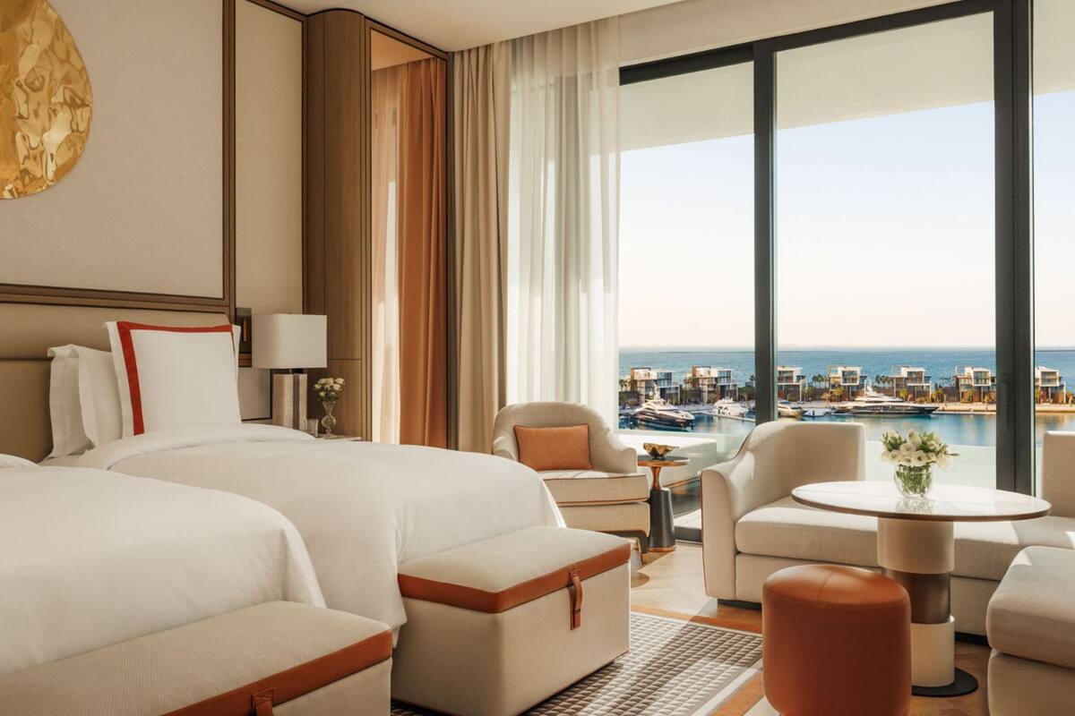 A hotel room with two beds, a sitting area with a view of the sea, and a balcony with a pool and boats.