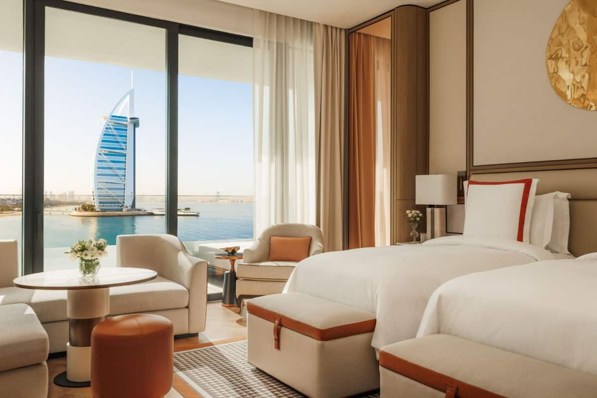 A hotel room with two beds, a sofa, a small table, and a view of a distinctive building by the water.