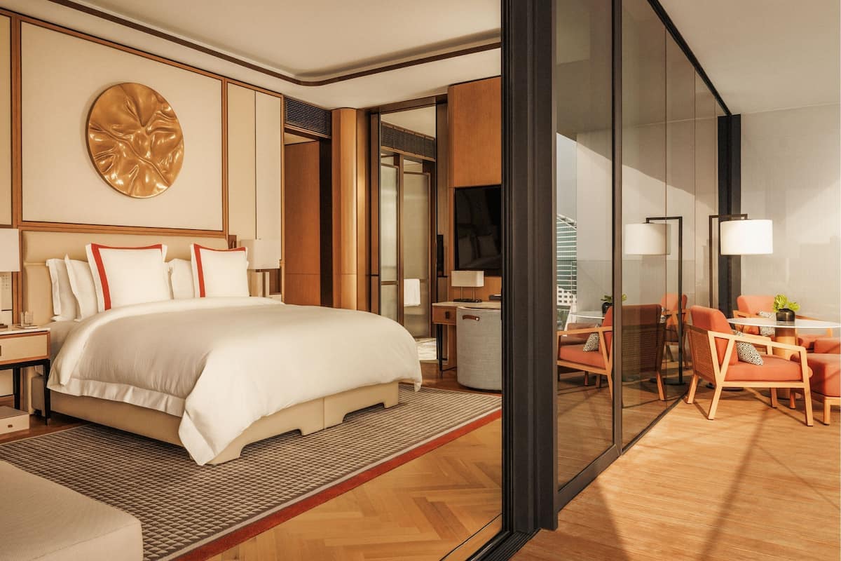 A modern hotel room with a large bed, a sitting area with orange chairs, a round decorative wall piece, and a large mirror.