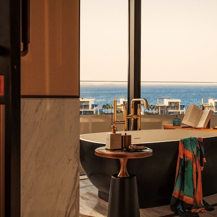 A modern bathroom with a large bathtub, a round mirror, and a view of the ocean.