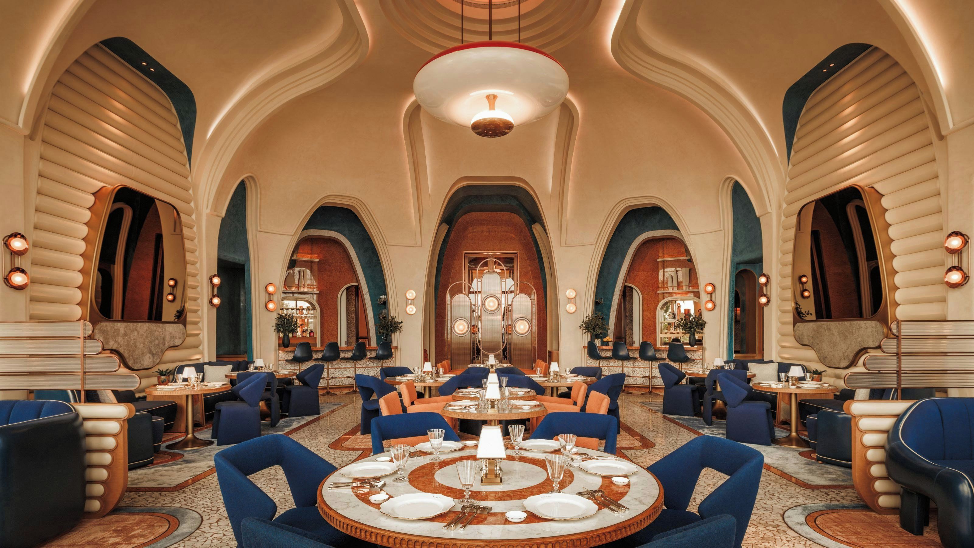 A spacious dining area with high ceilings and arched doorways, featuring round tables and blue upholstered chairs.