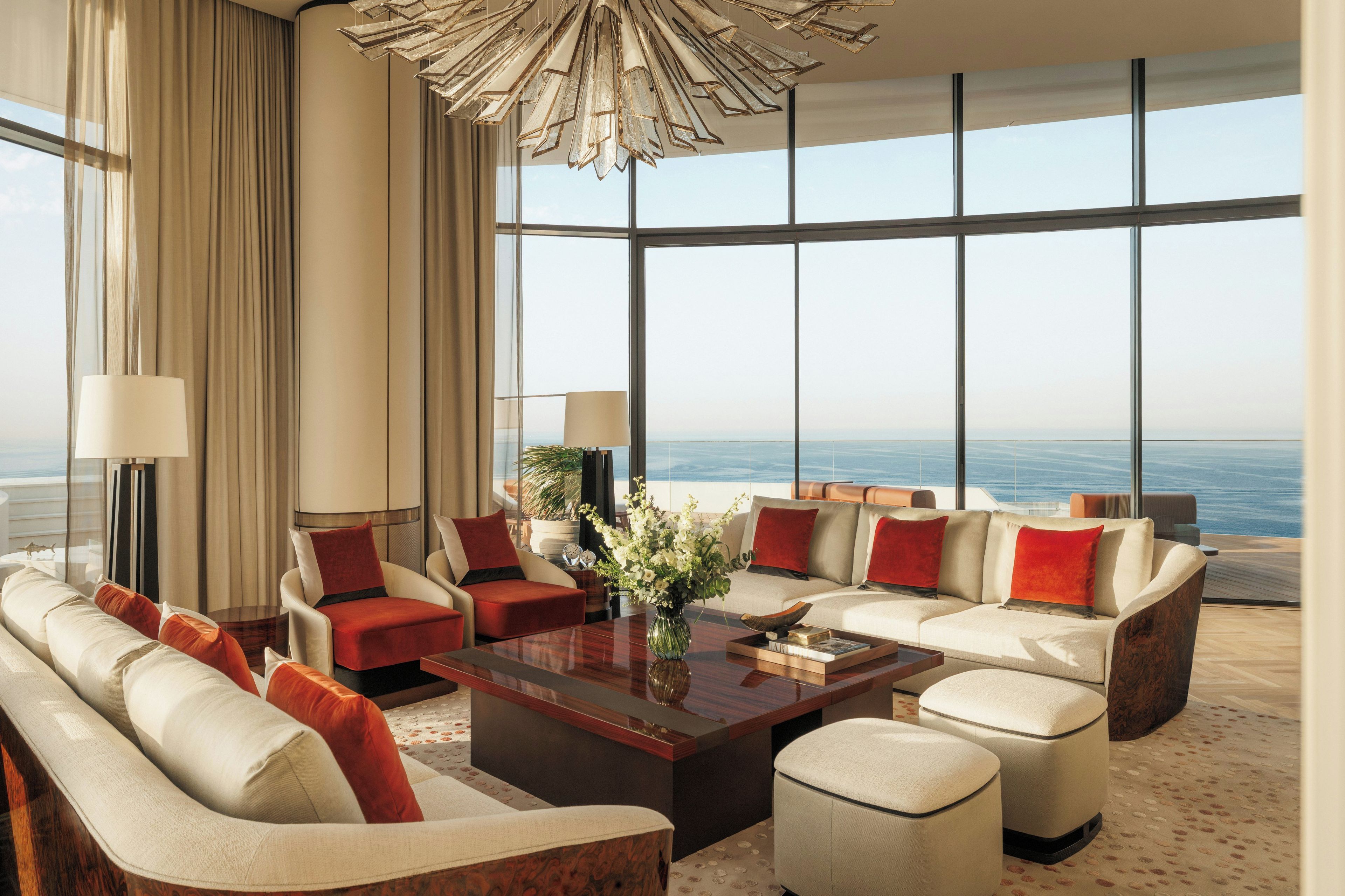Une chambre d’hôtel moderne dotée d’une grande fenêtre donnant sur la mer, d’un espace salon central avec une table basse et d’un lustre.