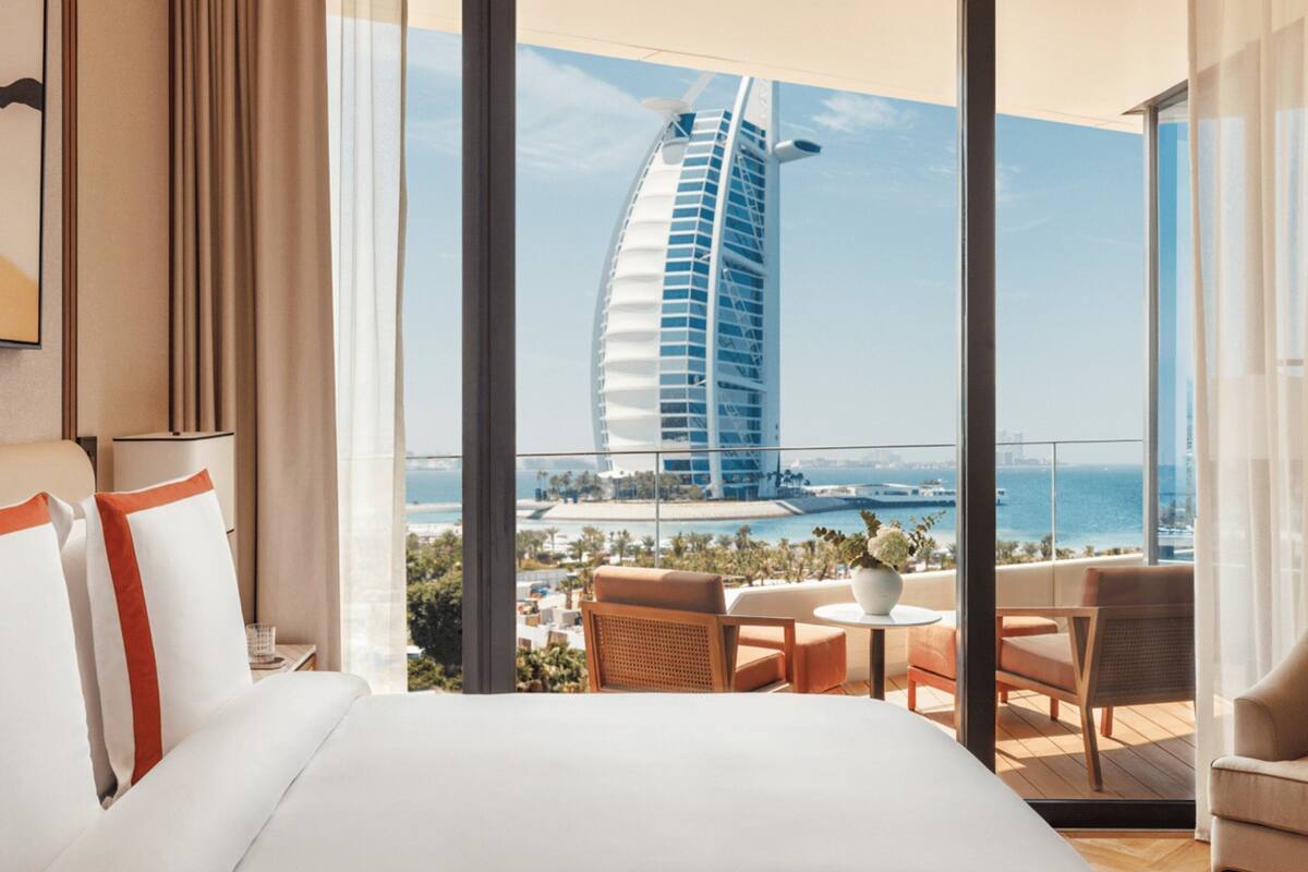 A hotel room with a large bed, a view of a distinctive sail-shaped building, and a balcony with seating.