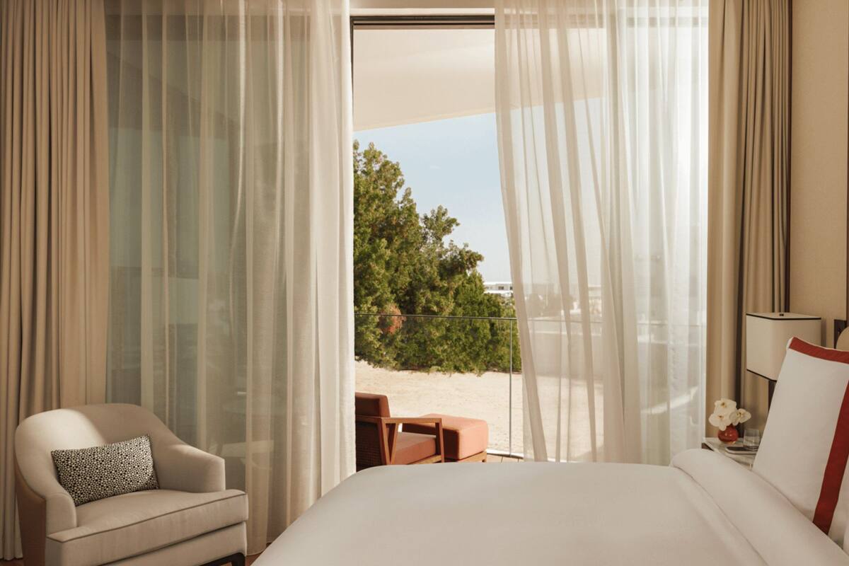 A hotel room with a bed, a chair, a small table, and a view of the outdoors.