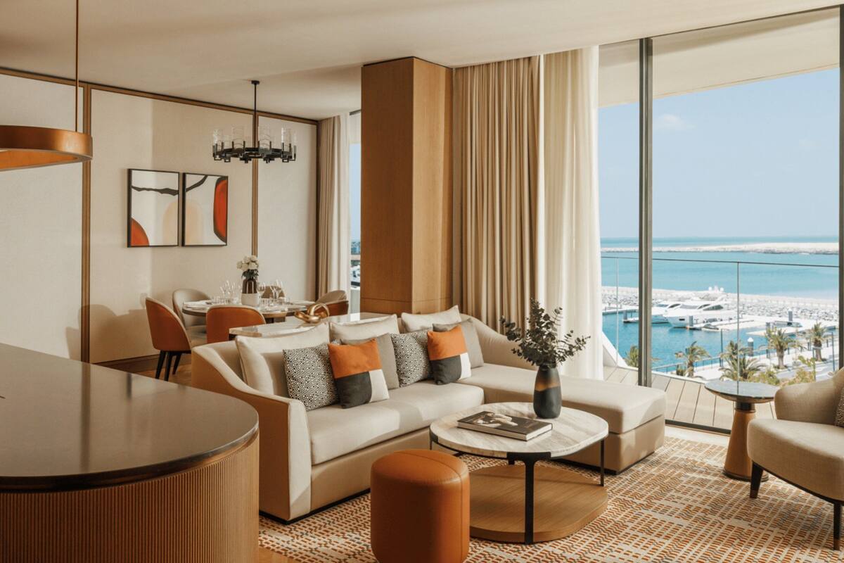 A modern living room with a sofa, coffee table, and chairs, a dining area with a table and chairs, a large window with a view of the sea, and a bar area.