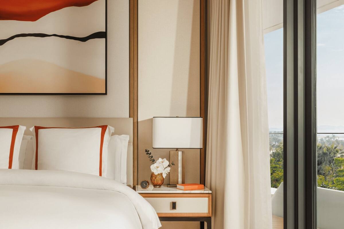 A hotel room with a large bed, a nightstand with a lamp, a vase with flowers, and a view of greenery through the window.