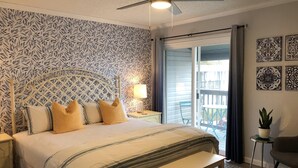 A bedroom with a bed, a bench, a nightstand, a lamp, a ceiling fan, a balcony with a chair, and decorative wall art. - Walk to beach. Perfect for families & couples. Pool & tennis onsite. (Fernandina Beach)