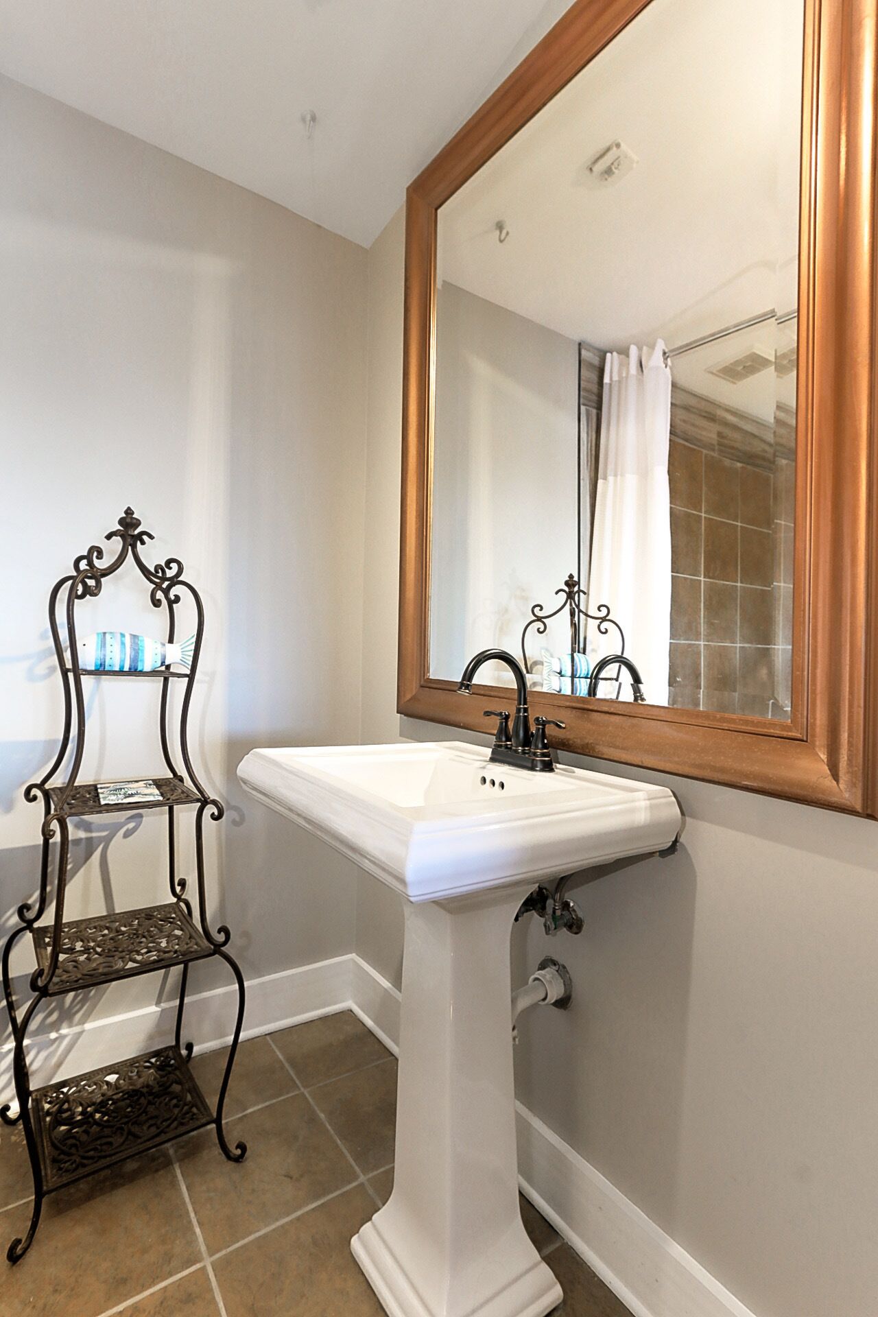 Une salle de bain avec un lavabo sur colonne blanc, un grand miroir avec un cadre en bois et une étagère en métal noir ornée d’objets décoratifs.