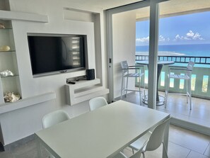A modern living room with a flat-screen TV, a dining table, and chairs. There is a balcony with a view of the ocean. - High floor King Bed beachfront access big balcony (Luquillo)