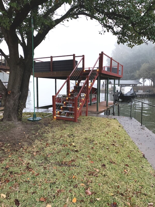 Une terrasse en bois avec un escalier menant à une plateforme d’observation surplombant l’eau, le tout entouré de pelouse et d’un arbre.