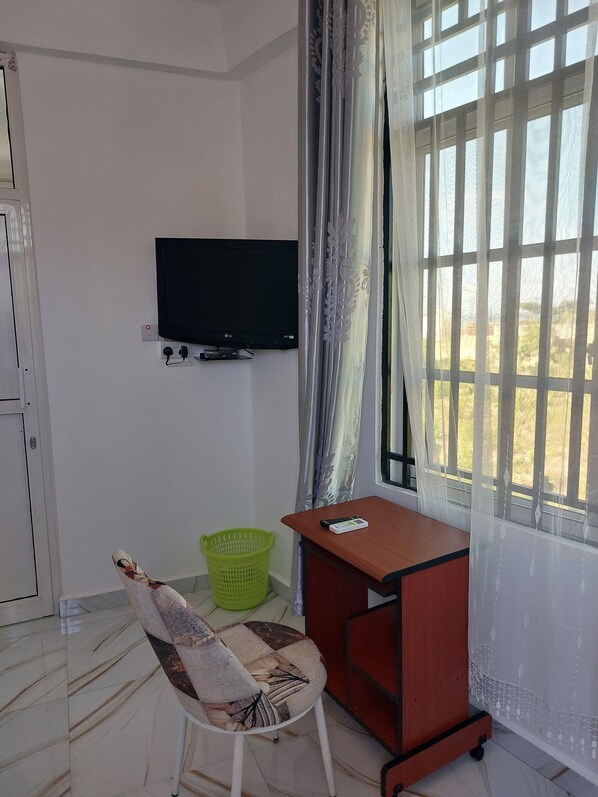 Une pièce avec un téléviseur fixé au mur, un bureau avec une chaise, un panier vert et une fenêtre avec des rideaux.