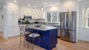 A modern kitchen with white cabinetry, a central island with bar stools, stainless steel appliances, and a large window. - Marshside Escape: Walk to Cockle Cove (South Chatham)