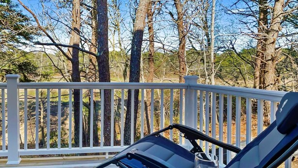 A deck with a white railing, a black lounge chair, and a green woven ottoman. - Marshside Escape: Walk to Cockle Cove (South Chatham)