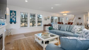 A spacious living room with a large sofa, a glass coffee table, and a fireplace. There are multiple windows providing natural light and views of the outdoors. - Marshside Escape: Walk to Cockle Cove (South Chatham)