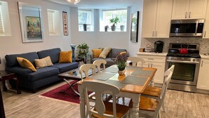 Un salon moderne avec un canapé bleu foncé, des chaises blanches et une table à manger en bois. On y trouve également un coin cuisine équipé d’appareils électroménagers en acier inoxydable et une bouilloire rouge posée sur la cuisinière.
