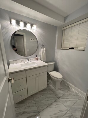 A bathroom with a white vanity, a round mirror, a toilet, and a window with blinds.