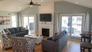 A spacious living room with a fireplace, a ceiling fan, and large windows. - Oceanside open concept home, roof deck with ocean and sunset bay views, firepit! (Beach Haven Crest)