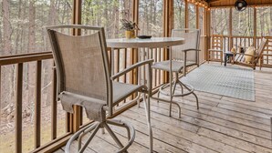 Un área de estar al aire libre, cubierta, con una mesa y sillas, rodeada de barandas de madera y ventanas grandes.